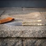 Damascus butterfly knife with skeletonised brass handles and a triquetra mark lying open on a granite step next to its soft tan leather carry pouch