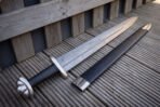 Damascus celtic knot panel viking sword and black scabbard angled across weathered decking, the interlaced knot band and polished steel throat catching low afternoon light