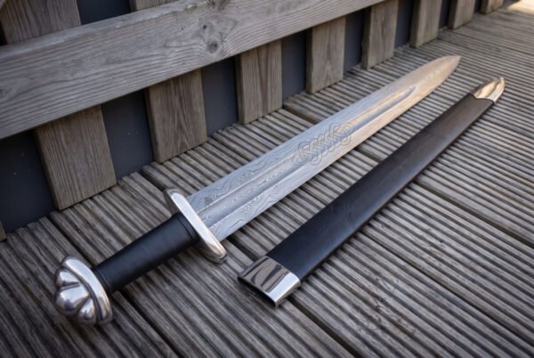 Damascus celtic knot panel viking sword and black scabbard angled across weathered decking, the interlaced knot band and polished steel throat catching low afternoon light