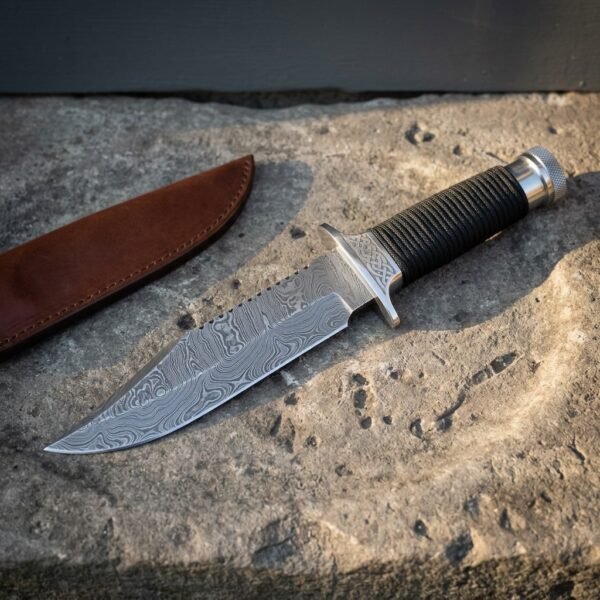 Damascus combat knife with a sawback spine, engraved steel guard and black cord wrapped grip resting on a pale limestone slab beside a brown leather sheath