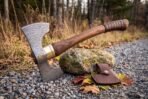 Damascus double bit axe leaning on a stone among autumn leaves on a gravel path, leather wrapped grip and brass collar