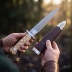 Damascus double edged dagger with carved Celtic knot bone handle held in two hands beside its burgundy leather sheath
