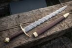 Damascus flamberge sword with a wavy pattern welded blade and brass ring guard lying on split timber beside its brown leather scabbard with brass mounts.