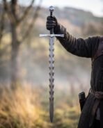 Damascus flame bladed sword held upright by the grip in a gloved hand with a misty valley behind