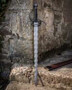Damascus flame bladed sword with an undulating edge and blackened swept hilt standing point down against a weathered stone wall beside a brown leather scabbard