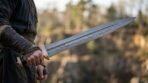 Damascus gladius short sword with bronze fittings held out at waist height by a man in dark clothes outdoors