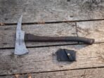 Damascus hatchet with rune engraved head and dark wood haft lying on grey boards beside a black leather blade mask