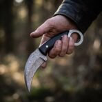 Damascus karambit knife with black handle and knotwork engraved steel finger ring held in a hand in the woods