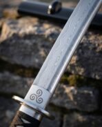 Damascus katana blade showing a wave pattern above a polished steel habaki engraved with a triskelion, seen close up