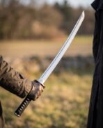 Damascus katana with black cord wrapped tsuka held in a gloved hand, curved blade raised against an open field