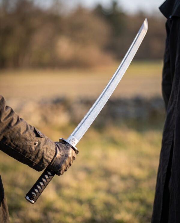 Damascus katana with black cord wrapped tsuka held in a gloved hand, curved blade raised against an open field