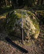 Damascus katana with black wrapped handle standing against a mossy boulder in the woods, black saya lying in front
