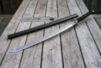 Damascus katana with black wrapped tsuka and steel tsuba lying across a grey timber table beside its saya