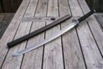 Damascus katana with visible temper line resting next to its black wooden saya on a weathered wooden table