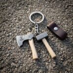 Damascus keyring charms shaped as a small axe and hammer on wooden handles with a steel ring, laid on gravel beside a leather sheath