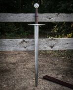Damascus knight sword standing point down in earth against a weathered fence, folded steel blade, brown leather grip and disc pommel engraved with a triquetra