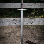Damascus knight sword standing point down in earth against a weathered fence, folded steel blade, brown leather grip and disc pommel engraved with a triquetra