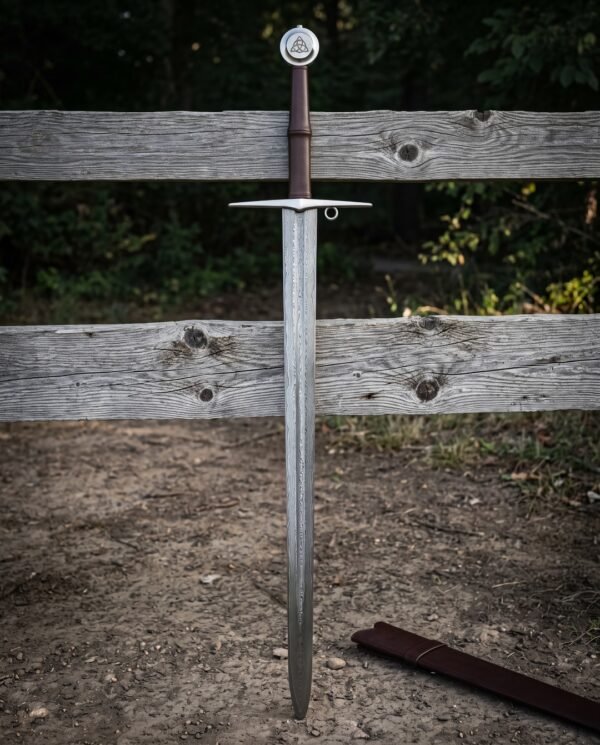 Damascus knight sword standing point down in earth against a weathered fence, folded steel blade, brown leather grip and disc pommel engraved with a triquetra