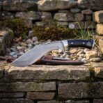 Damascus kukri with black handle scales, wider view resting on a dry stone wall with pebbles and the leather belt sheath