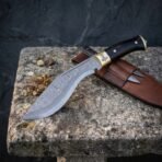 Damascus kukri with black horn grip and engraved brass bolster, slightly closer view on a stone bench with the companion knife sheath