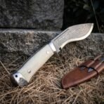 Damascus kukri with bone handle and wolf etched steel pommel, close up of the handle and pommel with the blade pointing upward