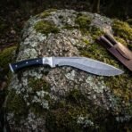 Damascus kukri knife with black buffalo horn handle, full length side profile laid flat on a large lichen covered boulder in the woods