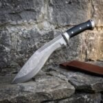 Damascus kukri knife with black micarta handle and engraved spine, angled on rough stone against an old stone wall with a leather sheath