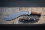 Damascus kukri knife with rosewood handle and steel bolster laid on a wooden beam above a black sheath with two small blades