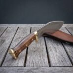Damascus kukri knife with walnut handle and brass fittings propped on grey wooden boards, sheath resting behind
