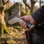 Damascus kukri knife with black handle and Valknut engraved steel butt cap held at the hip by a man with a leather belt sheath