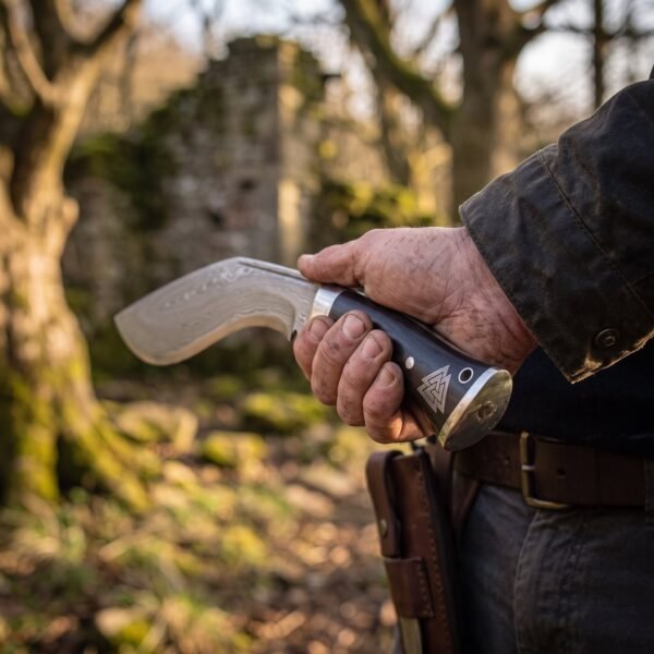 Damascus kukri knife with black handle and Valknut engraved steel butt cap held at the hip by a man with a leather belt sheath