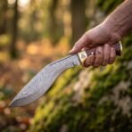 Damascus kukri knife with wooden handle and brass bolster engraved with a Valknut held in a hand in autumn woodland