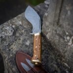 Damascus kukri with olive wood handle standing point up against weathered stone, with the leather sheath and companion blades below