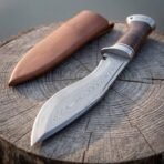 Damascus kukri with stacked leather grip and runed steel pommel, close angled view of blade and handle on a stump with tan sheath