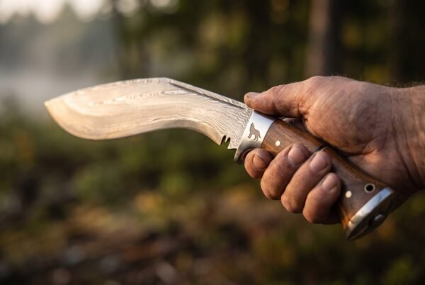 Damascus kukri with rosewood scales, steel bolster etched with a wolf and steel butt cap held in a hand in misty woods