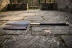 Damascus leaf blade spear with brass socket and dark shaft lying on old flagstones beside its brown leather blade cover
