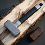 Damascus mjolnir hammer with a bold ladder pattern head, a knotwork engraved steel collar and a black leather wrapped haft, lying on split pale timber