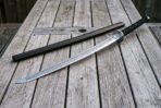 Damascus ninjato sword laid diagonally across weathered deck boards with its straight hardwood saya, showing the angled chisel point and triskele engraved steel guard