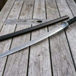 Damascus ninjato sword laid diagonally across weathered deck boards with its straight hardwood saya, showing the angled chisel point and triskele engraved steel guard