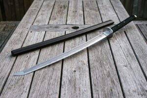 Damascus ninjato sword laid diagonally across weathered deck boards with its straight hardwood saya, showing the angled chisel point and triskele engraved steel guard