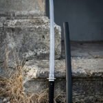 Damascus ninjato with a straight pattern welded blade, spiral engraved round guard and black cord handle standing beside its lacquered scabbard on stone steps