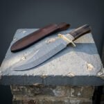 Damascus ram horn bowie knife with a brass guard and scrolled bolster resting on a pale stone plinth beside a stitched brown leather belt sheath
