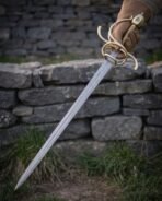 Damascus rapier with brass side rings and knuckle bow held in a gloved hand, point down against a stone wall