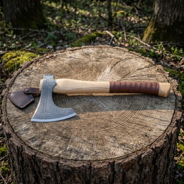 Damascus rune axe with a bearded head bordered in Elder Futhark, a pale ash haft and a leather grip wrap, resting on a cut tree stump in woodland