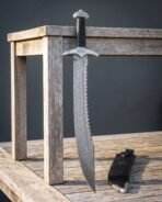 Damascus sawback sword with a serrated spine and curved knotwork guard standing against a weathered timber bench beside a black leather sheath with a steel chape