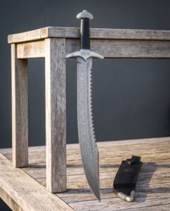 Damascus sawback sword with a serrated spine and curved knotwork guard standing against a weathered timber bench beside a black leather sheath with a steel chape