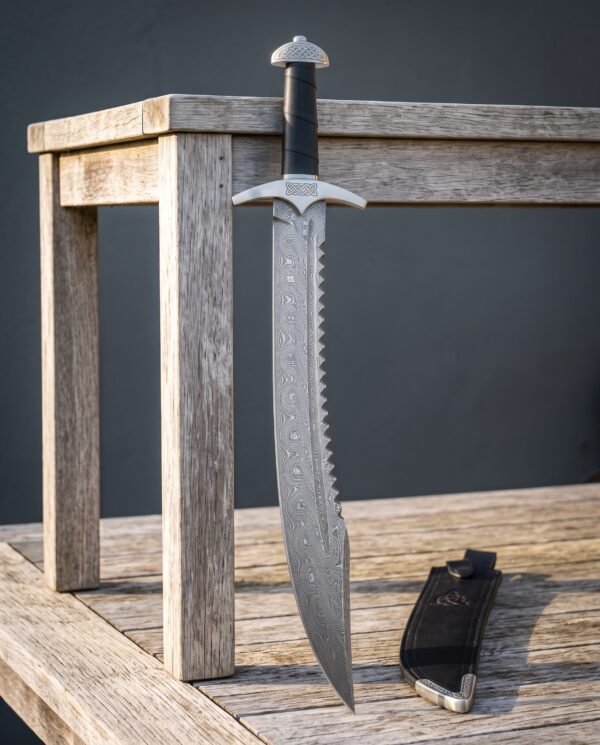 Damascus sawback sword with a serrated spine and curved knotwork guard standing against a weathered timber bench beside a black leather sheath with a steel chape