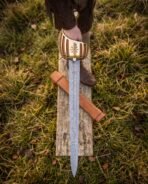 Damascus short sword with brass basket hilt and tan leather scabbard laid on a weathered board in the grass