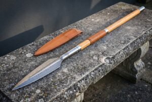 Damascus spear head on a pale ash haft with a leather wrapped grip resting on a weathered stone bench next to its tan leather blade sheath