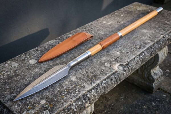 Damascus spear head on a pale ash haft with a leather wrapped grip resting on a weathered stone bench next to its tan leather blade sheath
